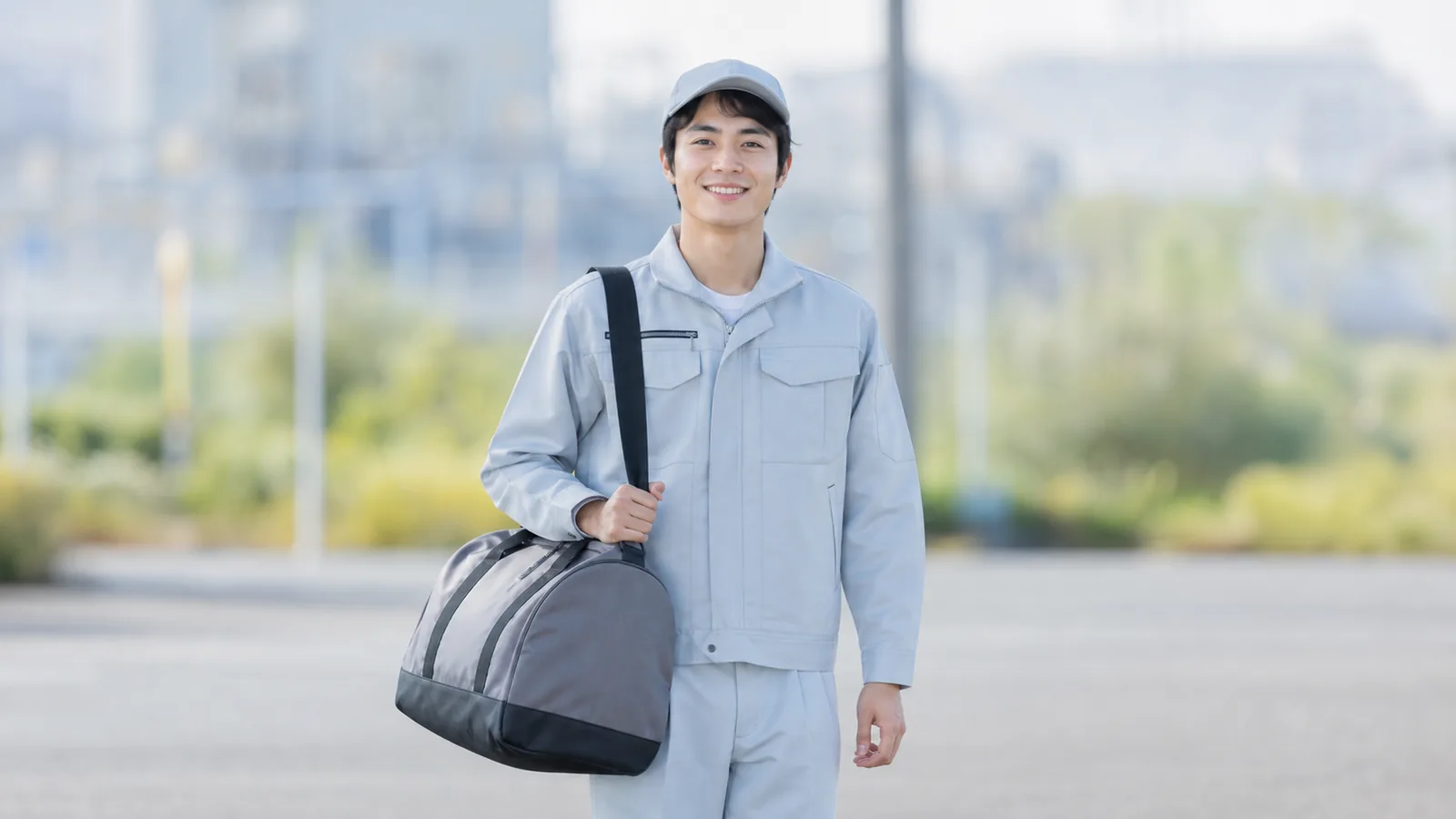 作業着で荷物ひとつを持ち、工場の前に立つスタッフのイメージ